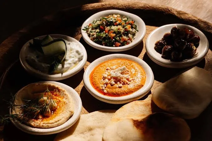 An assortment of Mediterranean dishes and dips served in small white bowls, including hummus topped with herbs, a tomato-based dip with crumbled cheese, a creamy white dip with cucumber garnish, a salad with chopped vegetables, and dark raisins, acco