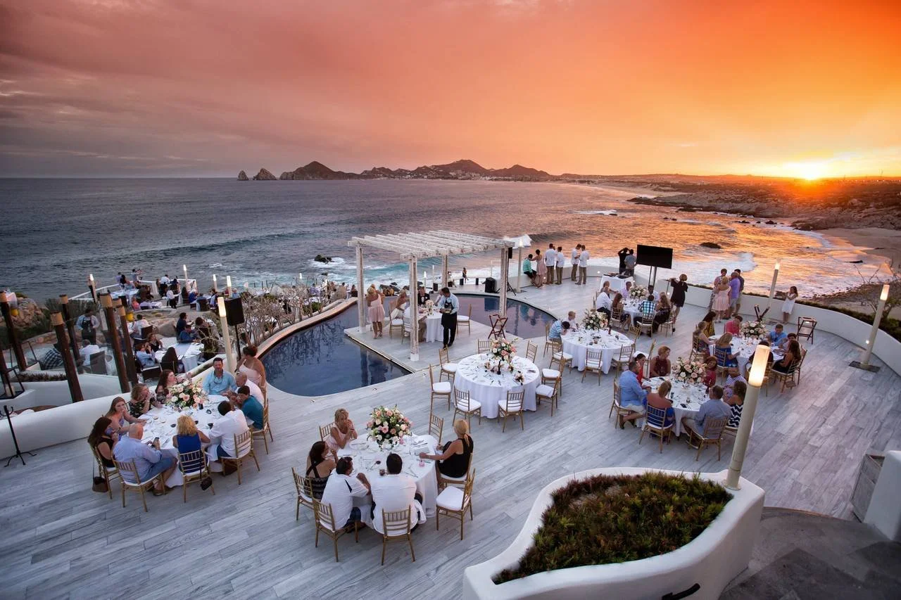 An outdoor event space on a terrace overlooking the ocean at sunset, with tables set for a dinner gathering and guests mingling.