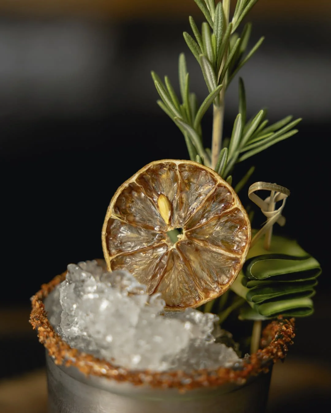 Close-up of a cocktail garnished with a dried lemon slice, fresh rosemary sprig, green leaves, and ice in a metal cup with a salted rim.
