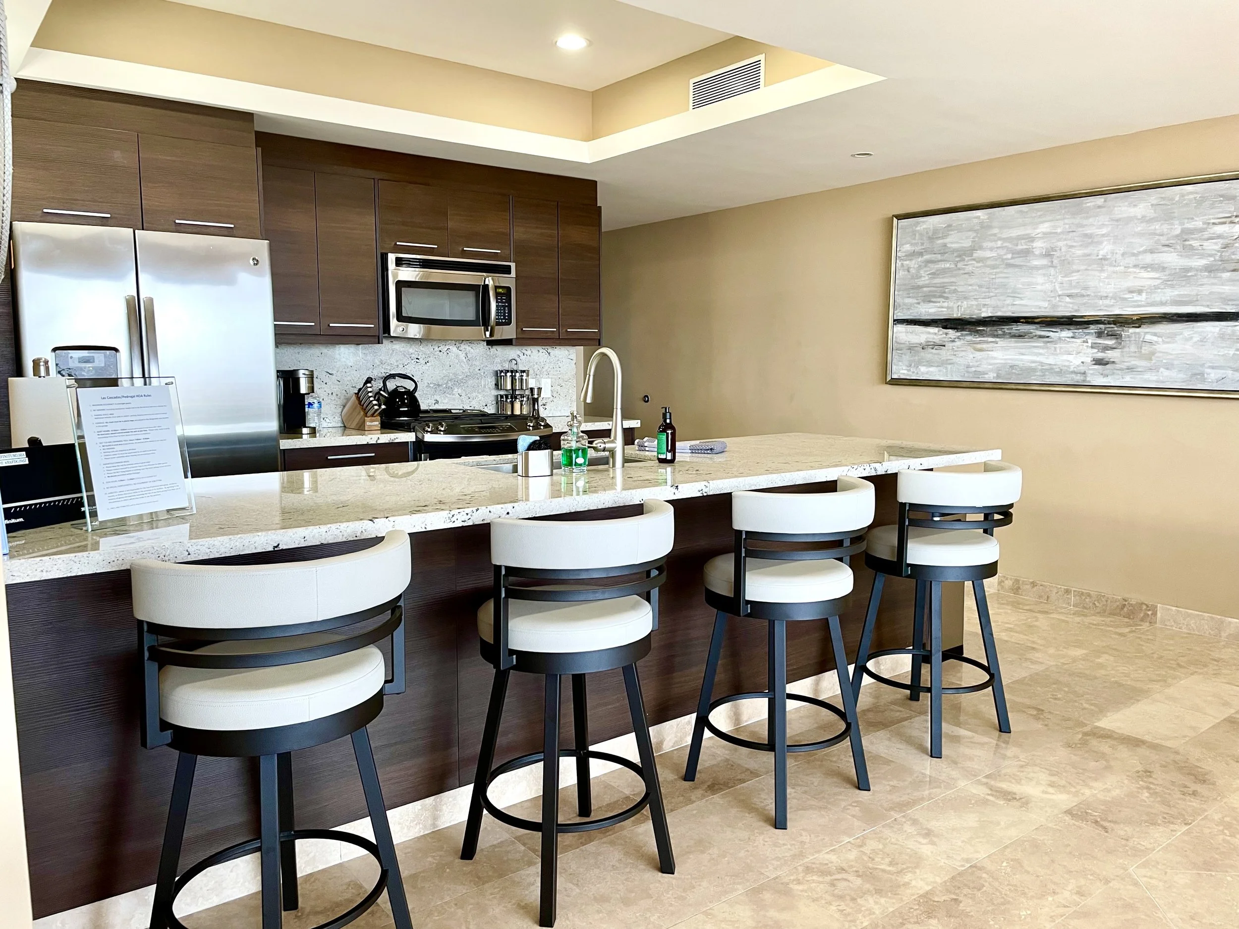 A modern kitchen with a beige and brown color scheme. It features a marble countertop island with four white and black bar stools, wooden cabinets, a microwave, and a stainless steel refrigerator. There is a large abstract painting on the wall and a sink with hand soap and a towel.