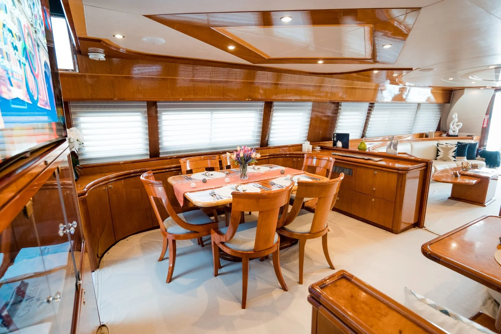 Interior of a dining area with a wooden dining table set with a pink tablecloth, plates, napkins, and a vase of flowers. Surrounding the table are wooden chairs with cushioned seats. Behind the table are several windows with white blinds, and to the 