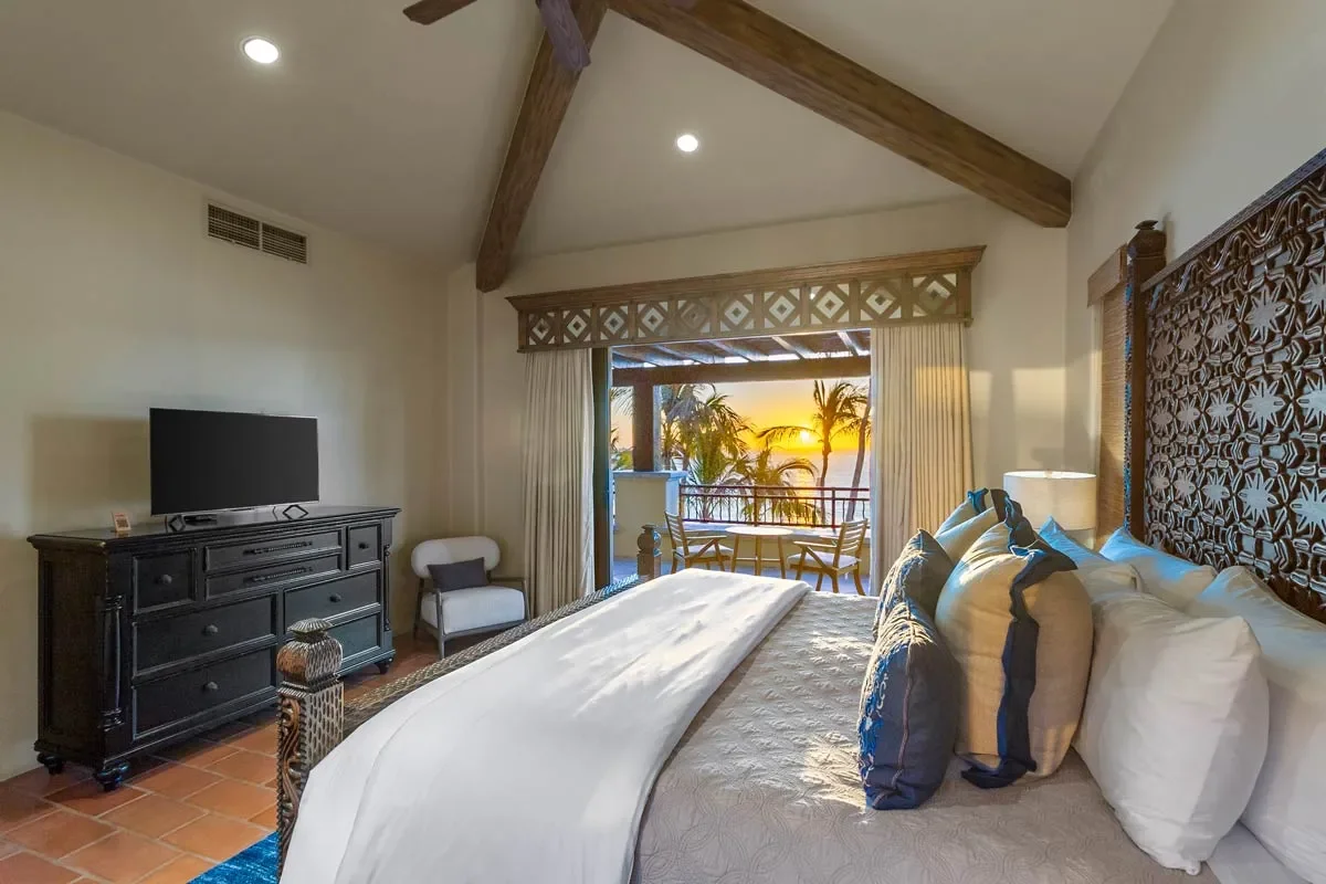 Hotel room with a large bed, decorative headboard, sitting area, and balcony with sunset view of palm trees.