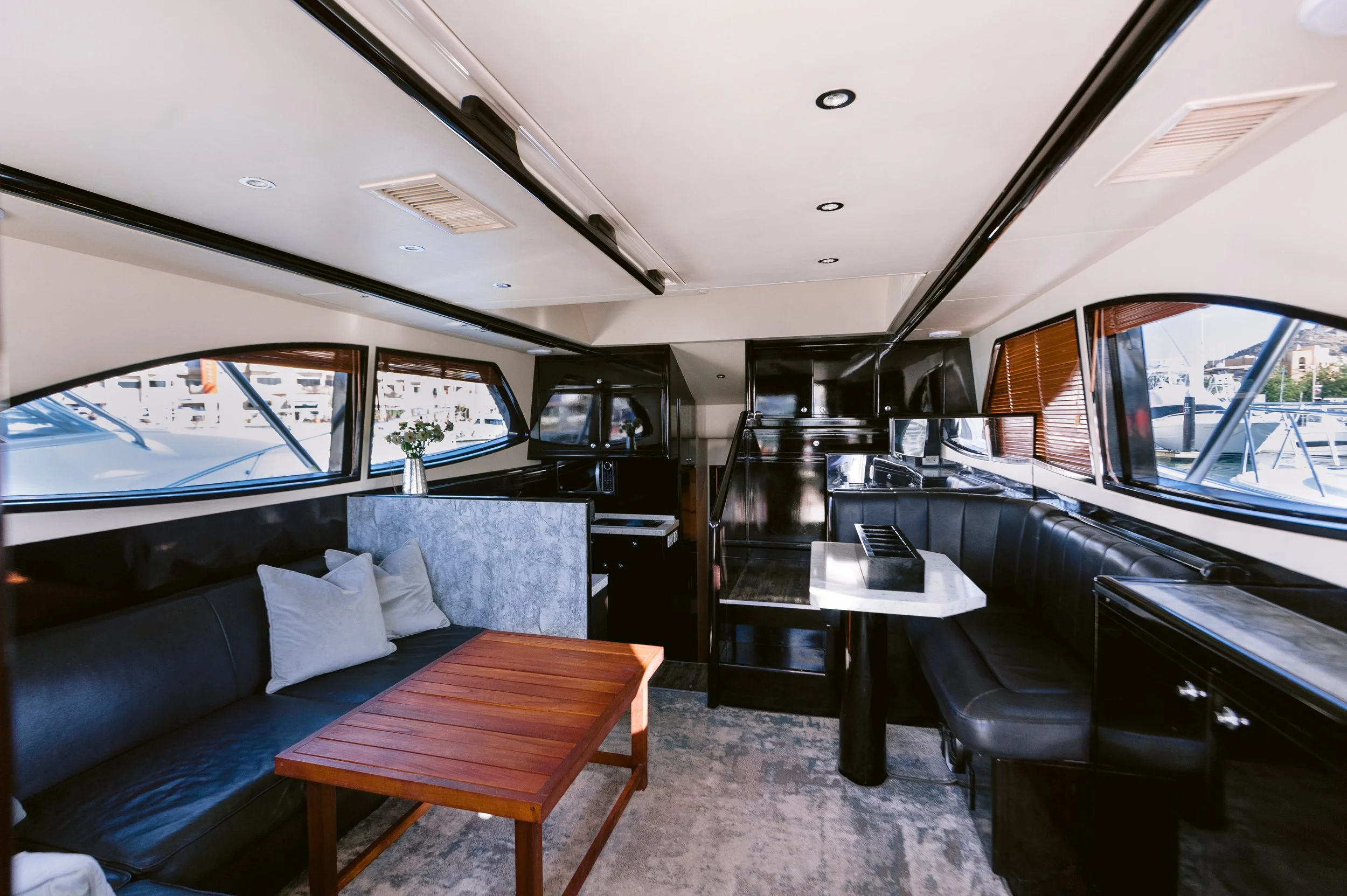Interior of a yacht with black leather sofas, a marble table, windows with blinds, and a view of boats docked outside.