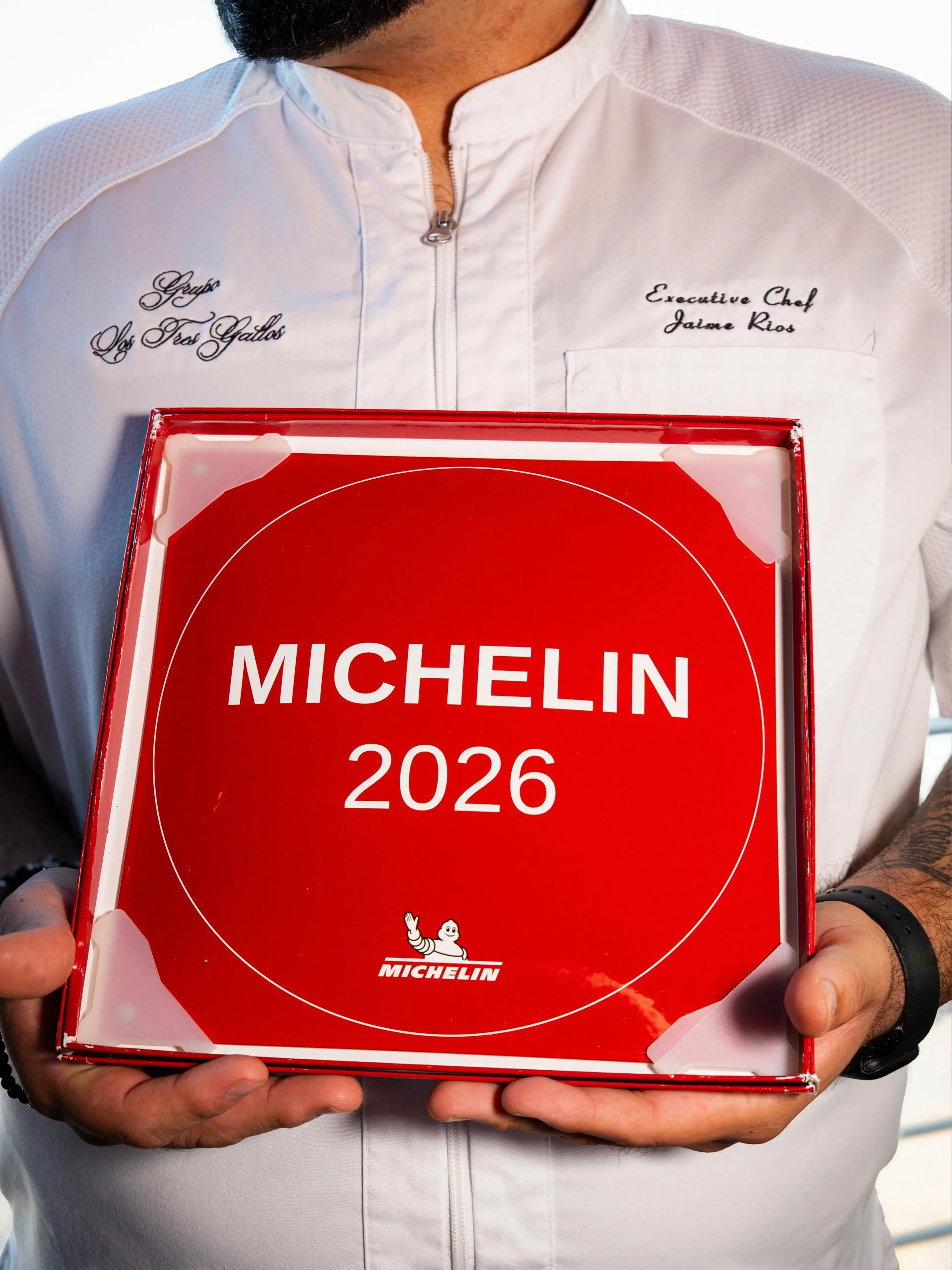 Person wearing a white jacket with embroidered text, holding a red Michelin guidebook with year 2026.