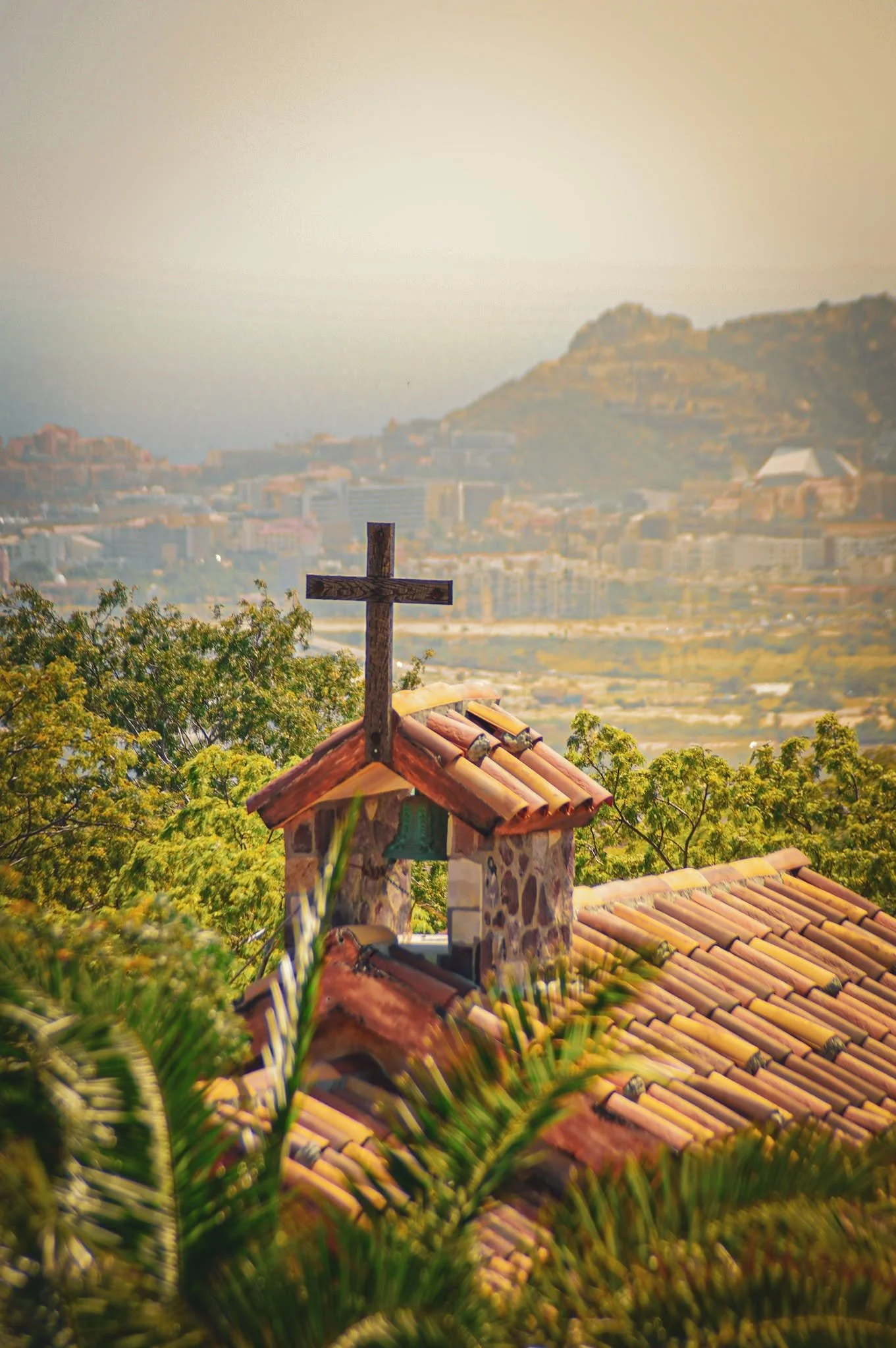 A wooden cross on top of a stone structure with a tiled roof, overlooking a cityscape with mountains in the background.