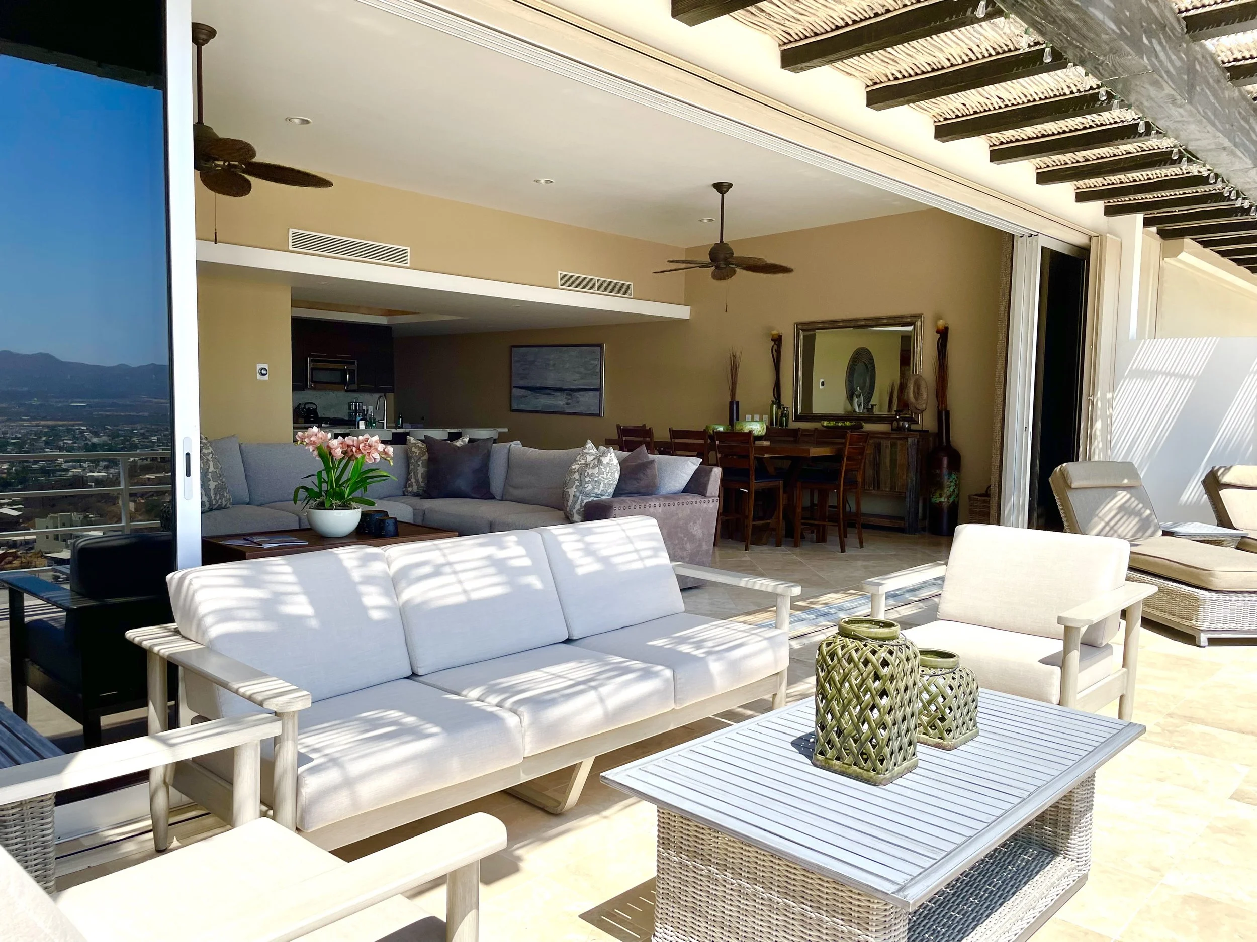 A spacious outdoor patio with white furniture, a potted pink flower, and decorative lanterns. The patio is shaded by a pergola with wooden beams and is connected to an indoor living space with a dining table and chairs. A cityscape and mountains are visible in the distance.
