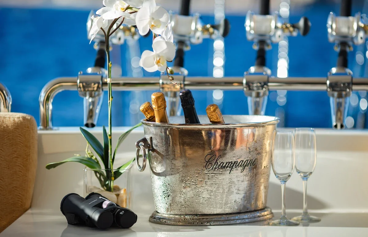 Bucket of champagne bottles with a flower and two glasses, set on a table near water
