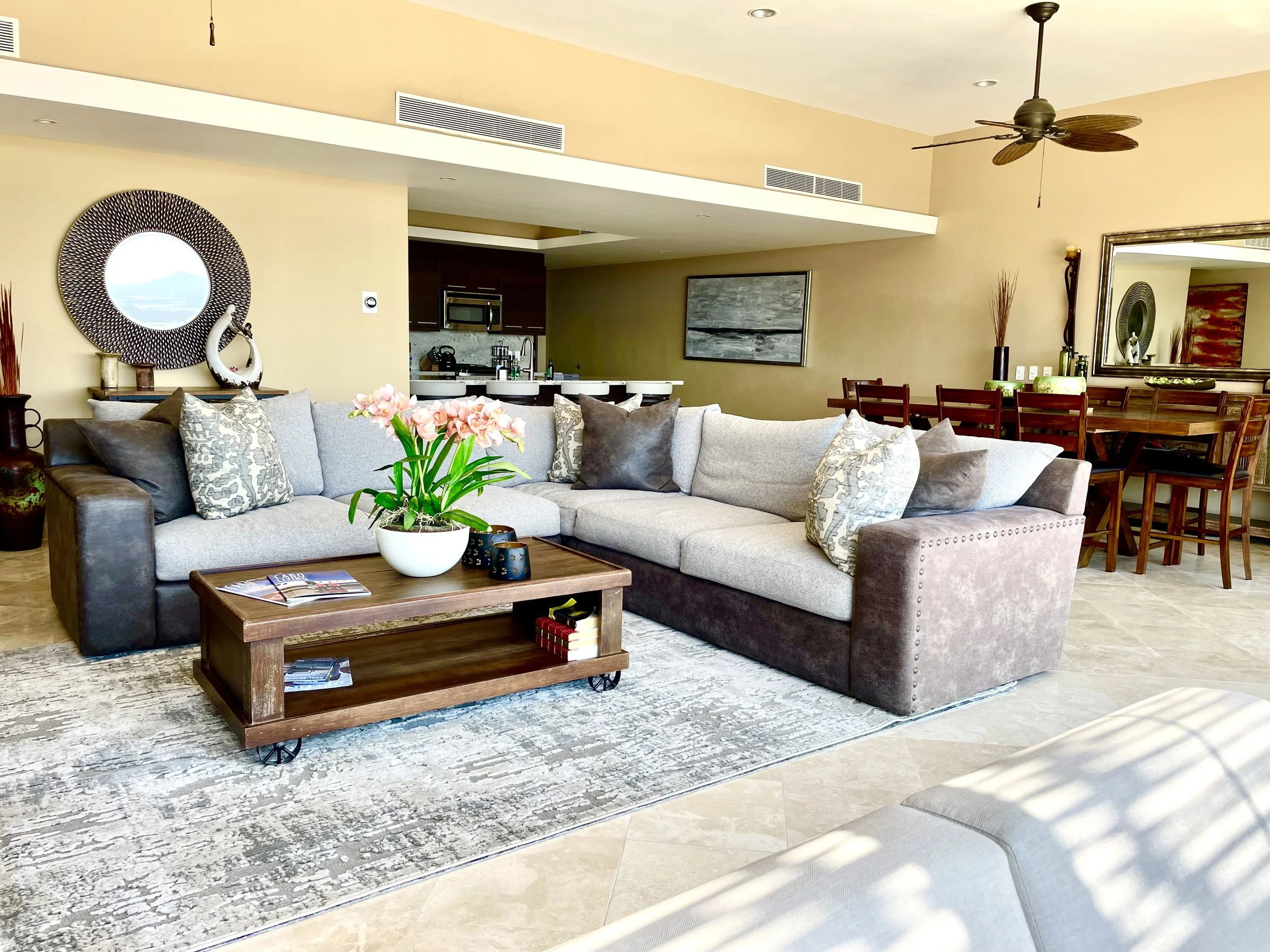 Living room with gray sectional sofa, wooden coffee table, decorative pillows, and pink flowers on the table. In the background, there is a dining area with a wooden table and chairs, a mirror, and wall decorations. The open kitchen is visible with dark cabinets, a microwave, and bar stools. Ceiling fans and air vents are also seen.