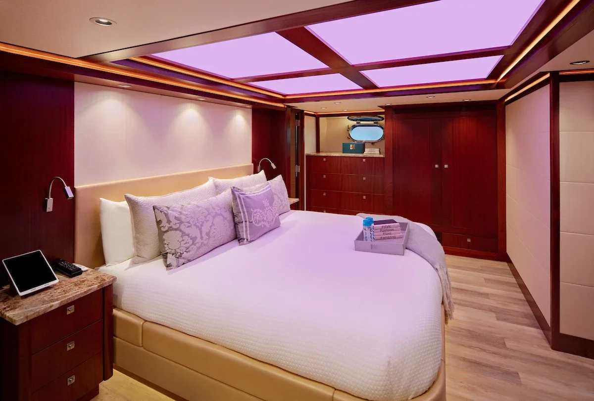 A luxurious bedroom with a large bed featuring decorative pillows, a wooden nightstand with a tablet and remote, and a tray with toiletries. The room has wood paneling, a padded wall, and a ceiling with pinkish-purple lighting panels.