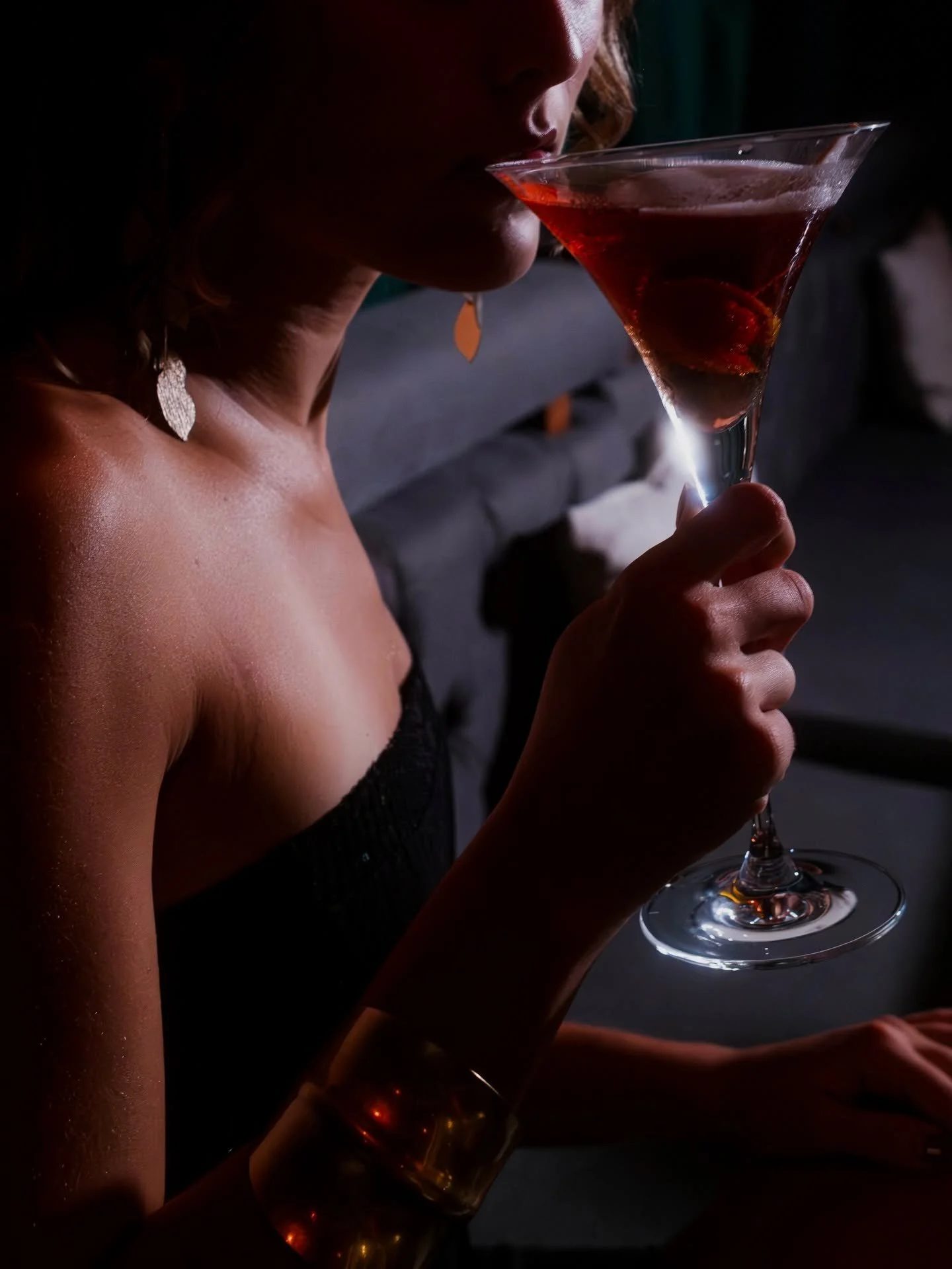 Person drinking a red cocktail from a martini glass, with part of their face and shoulder visible in a dimly lit setting.