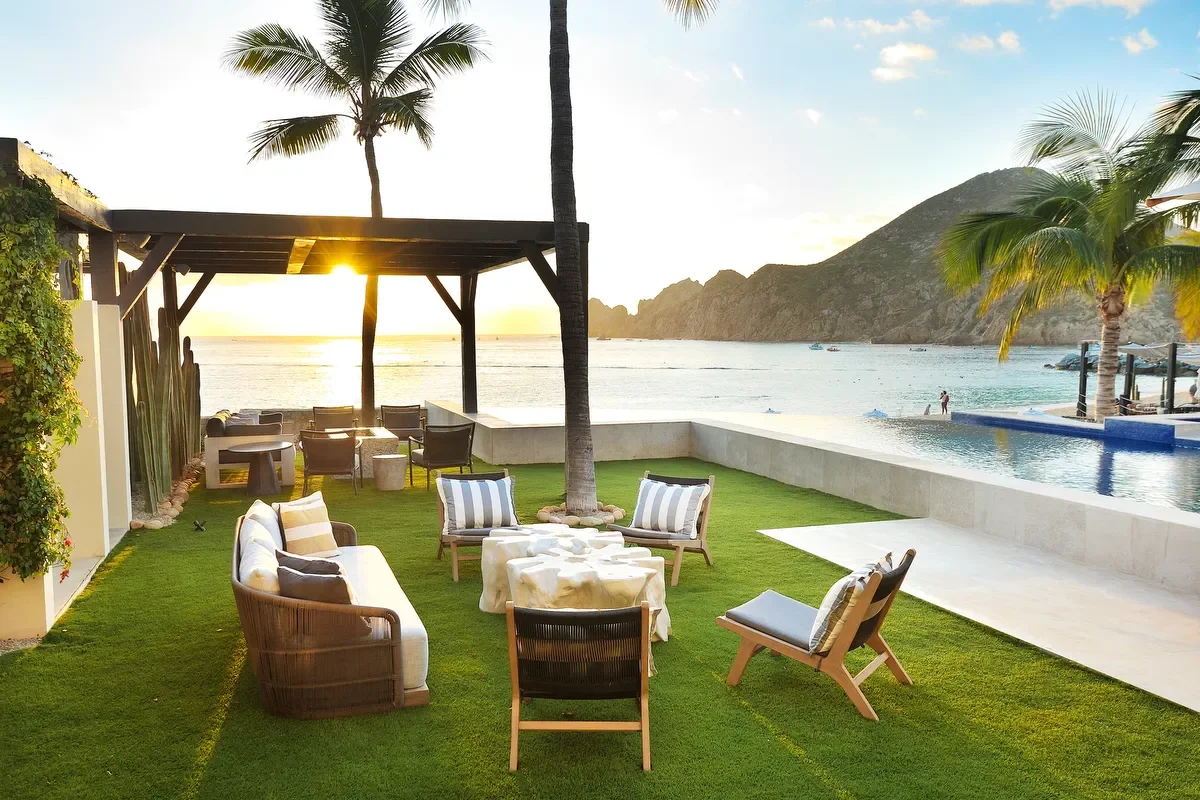 Outdoor seating area with sofas and chairs on a grassy lawn near a pool, overlooking an ocean with mountains in the background, during sunset.