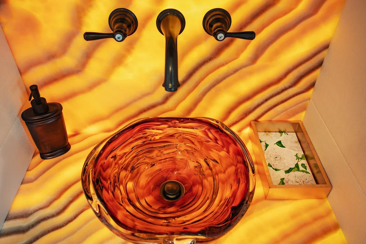 A bathroom sink with a glass basin, orange and swirled design, mounted on a backlit yellow and orange marble wall. A black faucet above, a soap dispenser to the left, and a wooden tray with white flowers to the right.