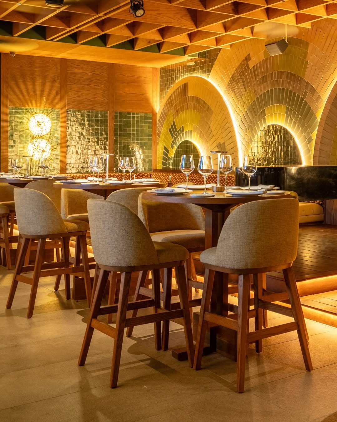 Elegant restaurant with round tables set with wine glasses, plates, and napkins. Wooden chairs and a decorative background featuring arched brick walls with warm lighting.