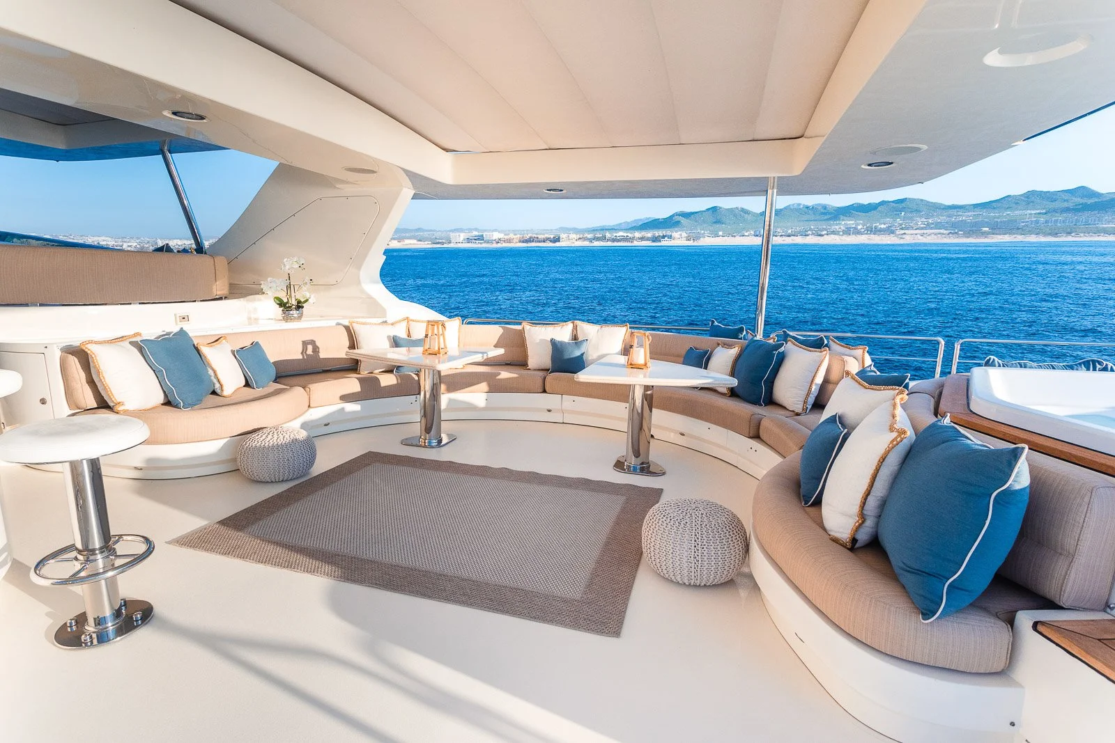 Luxury yacht deck with cushioned seating area, blue and white pillows, small tables, a rug, potted plant, and ocean view with distant mountains.