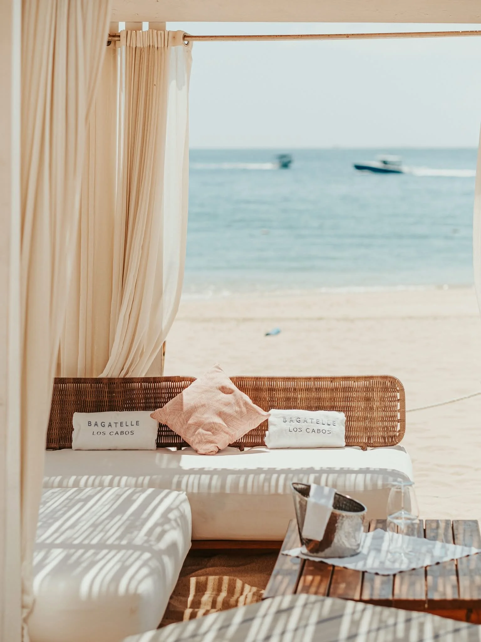Beachside lounge area with two white towels embroidered with "Bagatelle Los Cabos" on a wicker bench, a pink throw pillow, and a glass of white wine on a table in front. The background shows the ocean with boats and a sandy beach, seen through an open cabana with cream curtains.