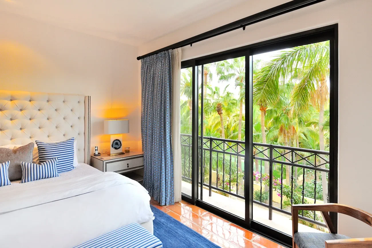 A bedroom with a large sliding glass door opening to a balcony overlooking lush green palm trees.