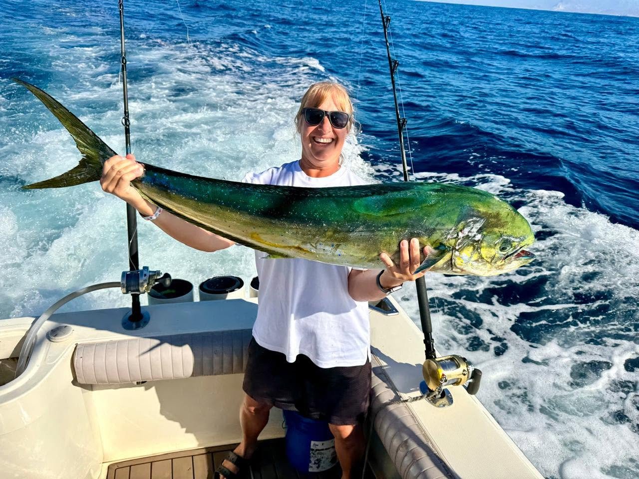 Woman on a boat holding a large fish, smiling, with the ocean in the background.