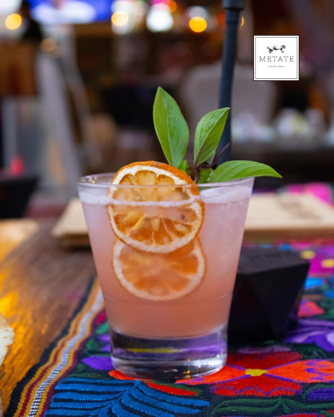 A pink cocktail garnished with slices of dried citrus, fresh green leaves, resting on a colorful embroidered tablecloth with a blurred background.