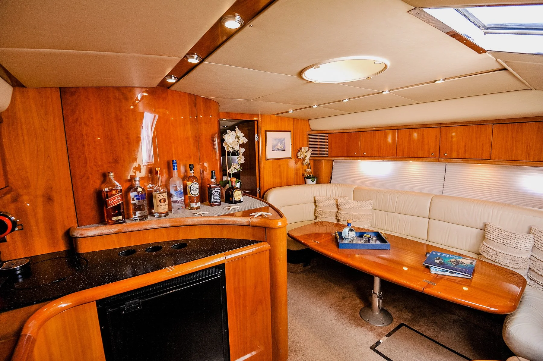 Interior of a boat or yacht with a wood-paneled ceiling and walls, a leather seating area with pillows, a wooden table with magazines, a built-in bar with liquor bottles, and a skylight overhead.