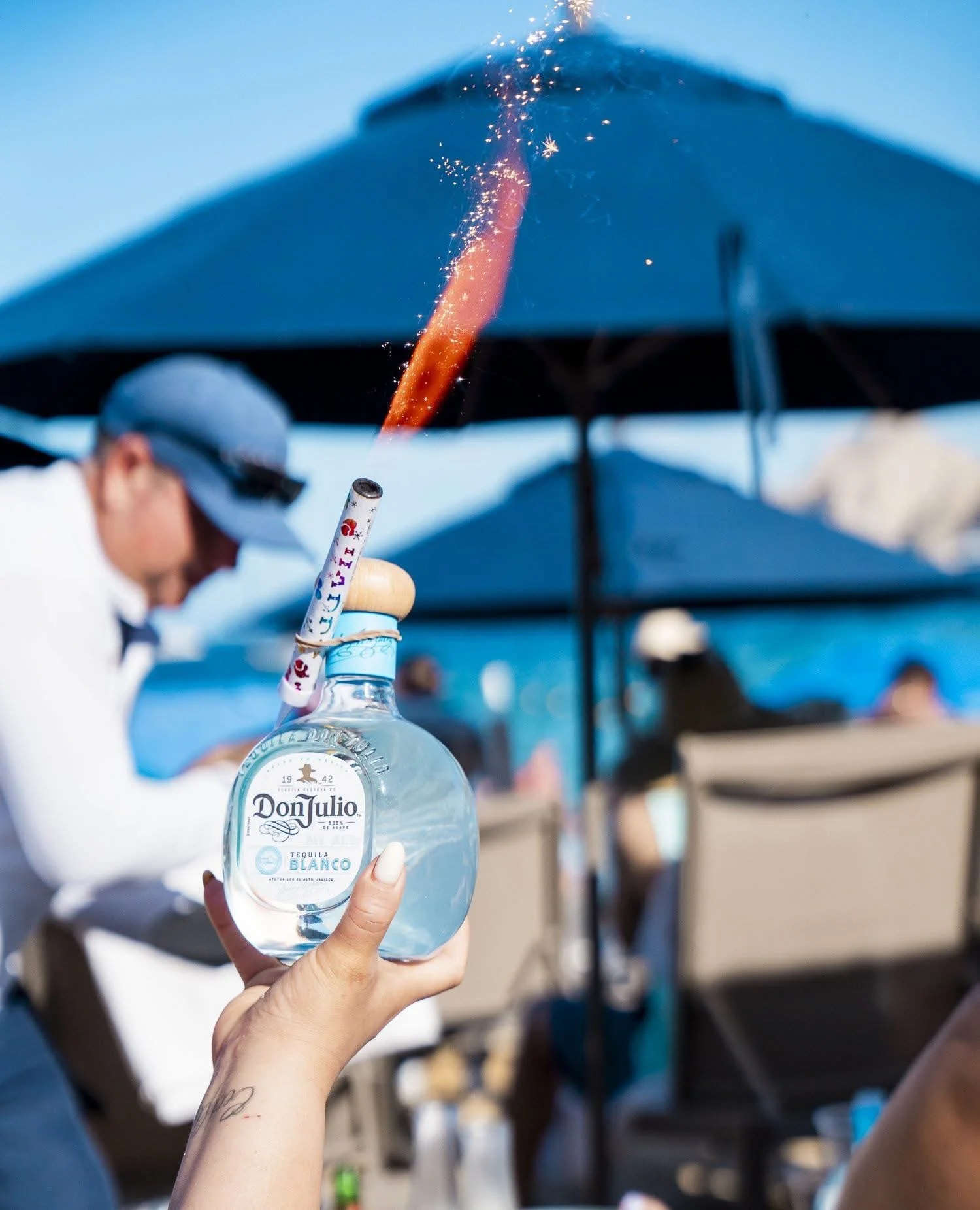A person holding a bottle of Don Julio tequila with a sparkler inserted into it, igniting a spark and flame, at an outdoor gathering with people and umbrellas visible in the background.
