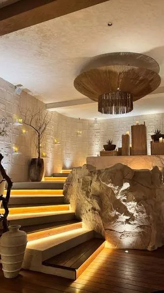 Elegant interior space with staircase illuminated by warm lighting, large rock feature, potted plants, and decorative elements on a curved wall.