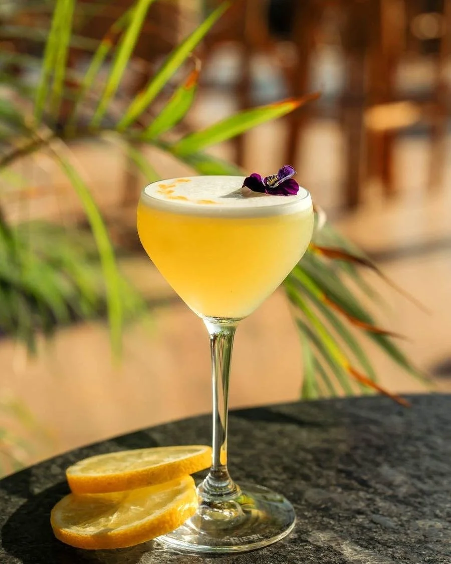 A yellow cocktail in a coupe glass garnished with purple edible flowers, with lemon slices on the table, set outdoors with blurred background greenery.