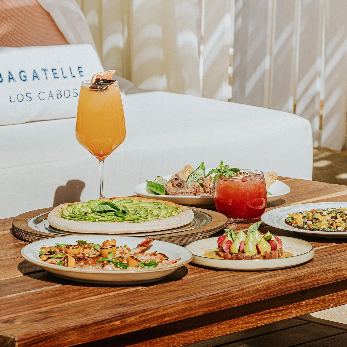 Assorted dishes and drinks on a wooden table, including a pizza with avocado slices, a seafood dish, a colorful salad, a watermelon and fruit dessert, and two cocktails, one orange and one red.
