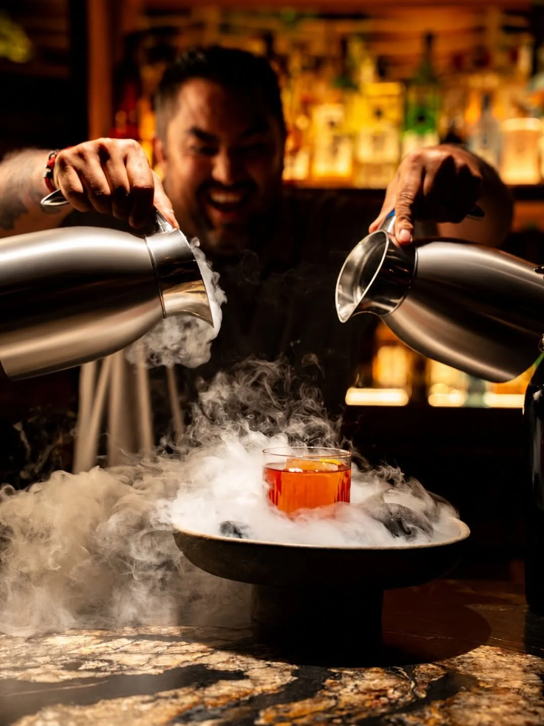 Bartender in a bar making a drink with dry ice, creating fog around a glass of red-colored beverage on a plate.