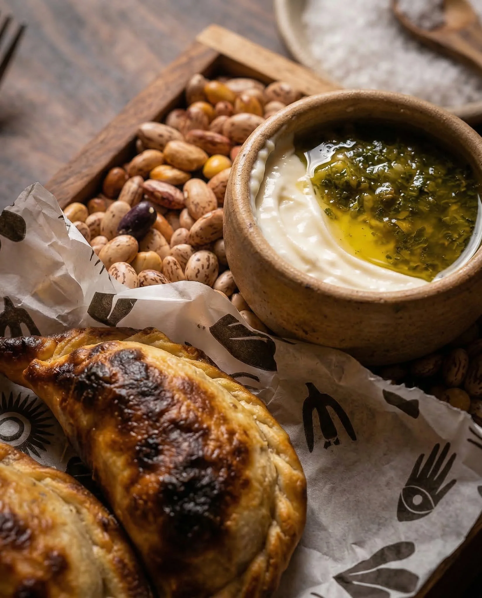 A wooden bowl with a creamy dip topped with herbs or pesto, surrounded by pinto beans, on parchment paper, with a grilled chicken piece in the foreground.