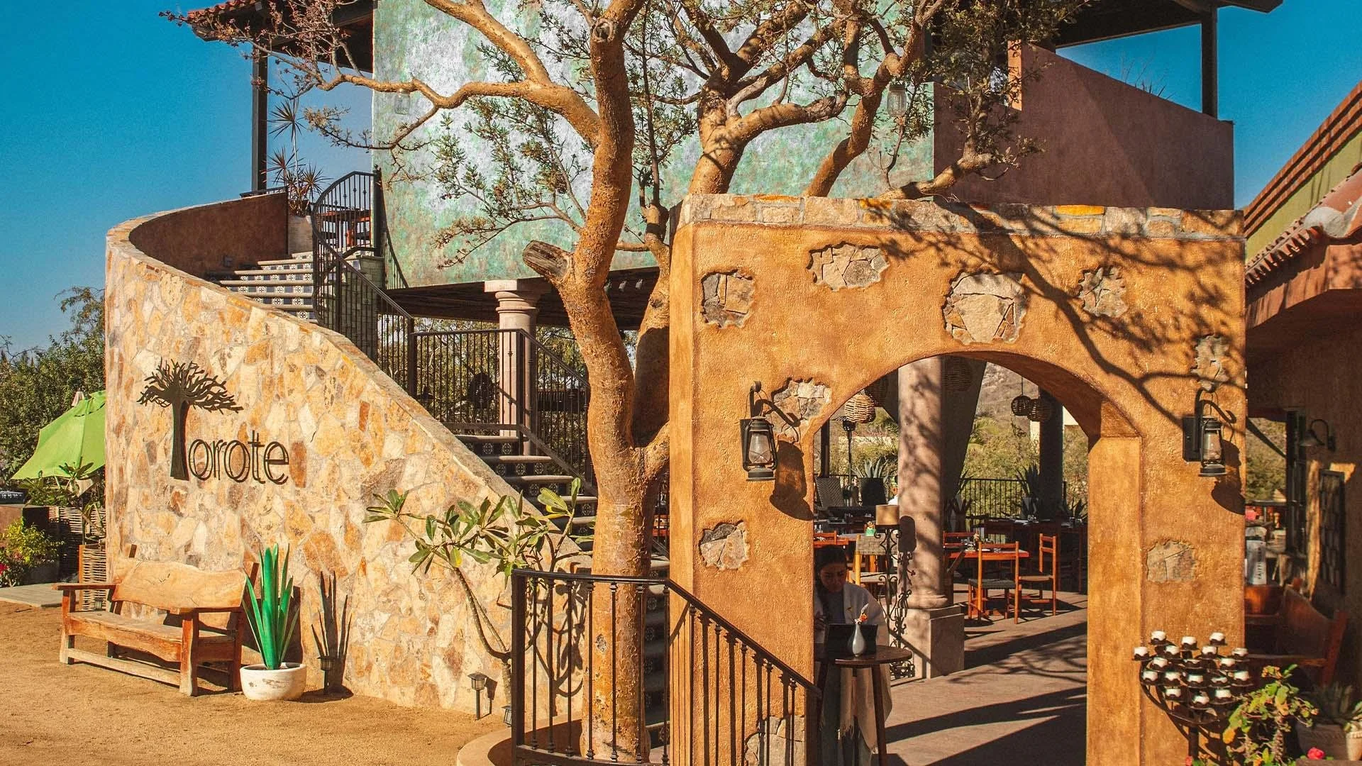Outdoor restaurant or cafe area with stone walls, arches, and a large tree. The word "Loret" is on the stone wall. There are tables and chairs, lantern-style lights, and decorative plants.