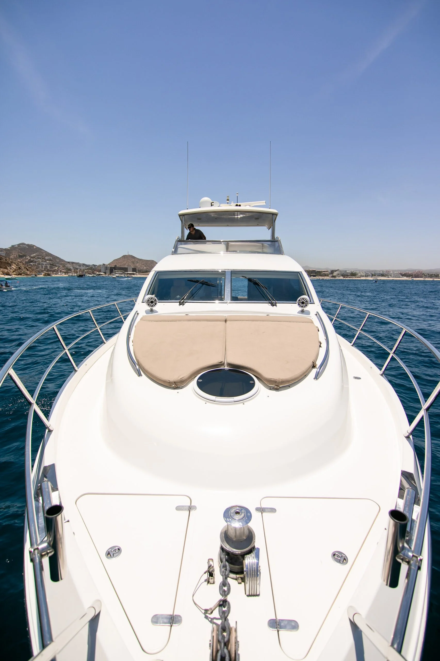 Front view of a white yacht on the water with a person on the upper deck, city buildings and hills in the background under a blue sky.