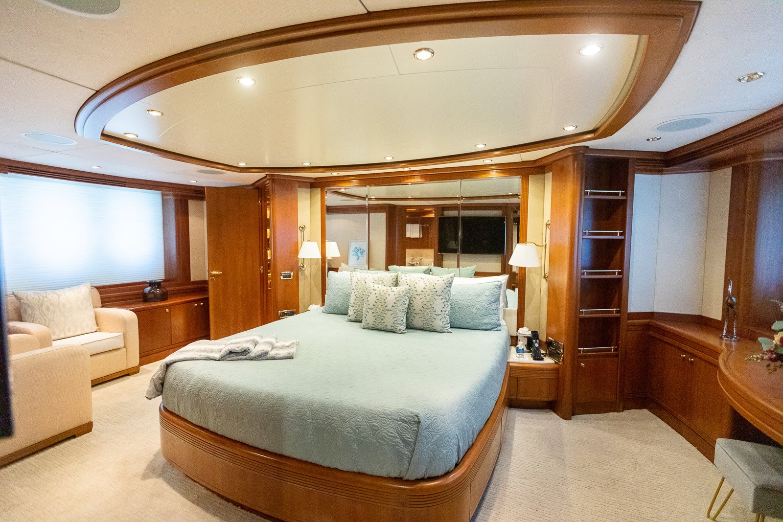 A luxurious bedroom with a large bed featuring light blue bedding and multiple pillows. The room has wooden walls and trim, a mirrored headboard, a sitting area with a cream armchair, and built-in shelving. There are wall-mounted lamps on either side