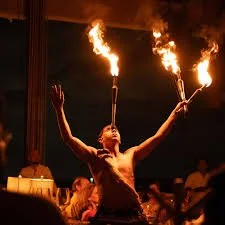 A street performer in a dark setting, juggling fire torches with flames on both hands, while people watch in the background.