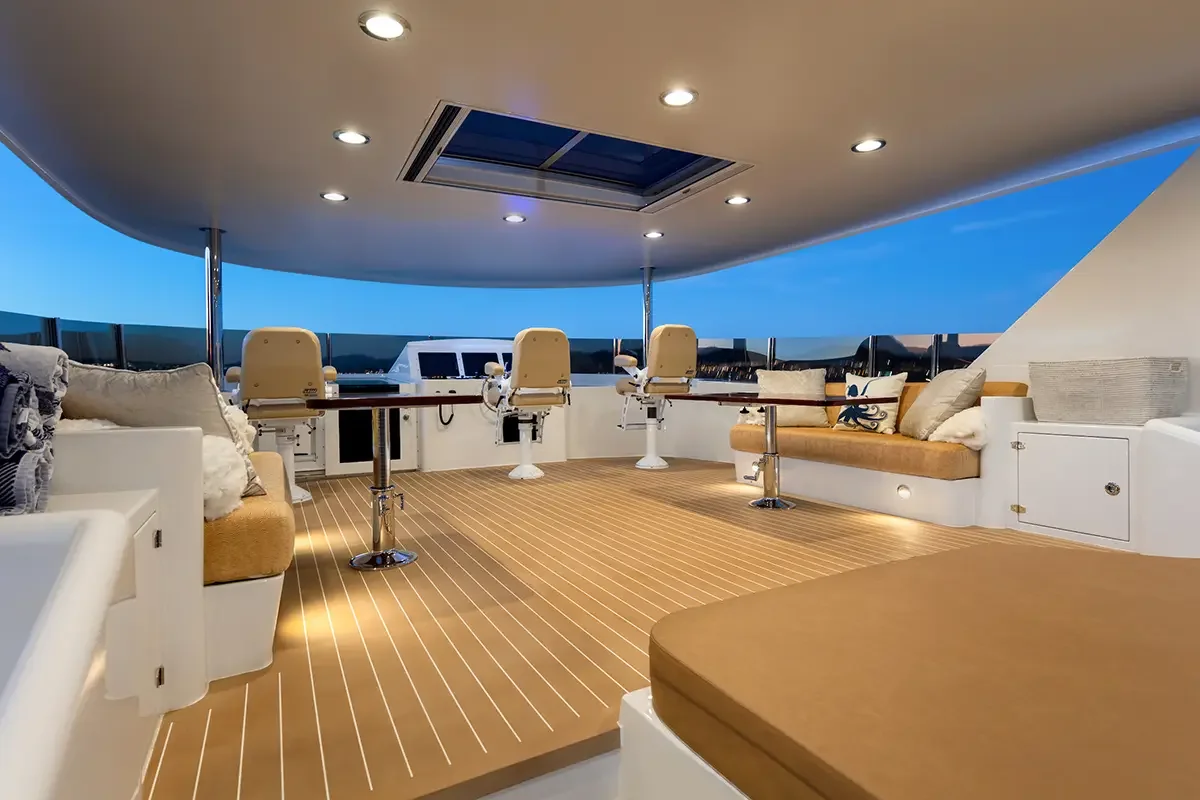 Outdoor yacht deck with seating, cushions, and a table, overlooking the ocean during twilight, with a skylight and overhead lights.