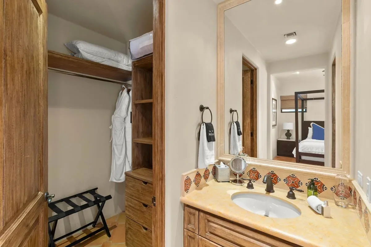 Bathroom vanity with a mirror, soap dispenser, and towels. Reflection shows a bedroom with a bed and nightstand.