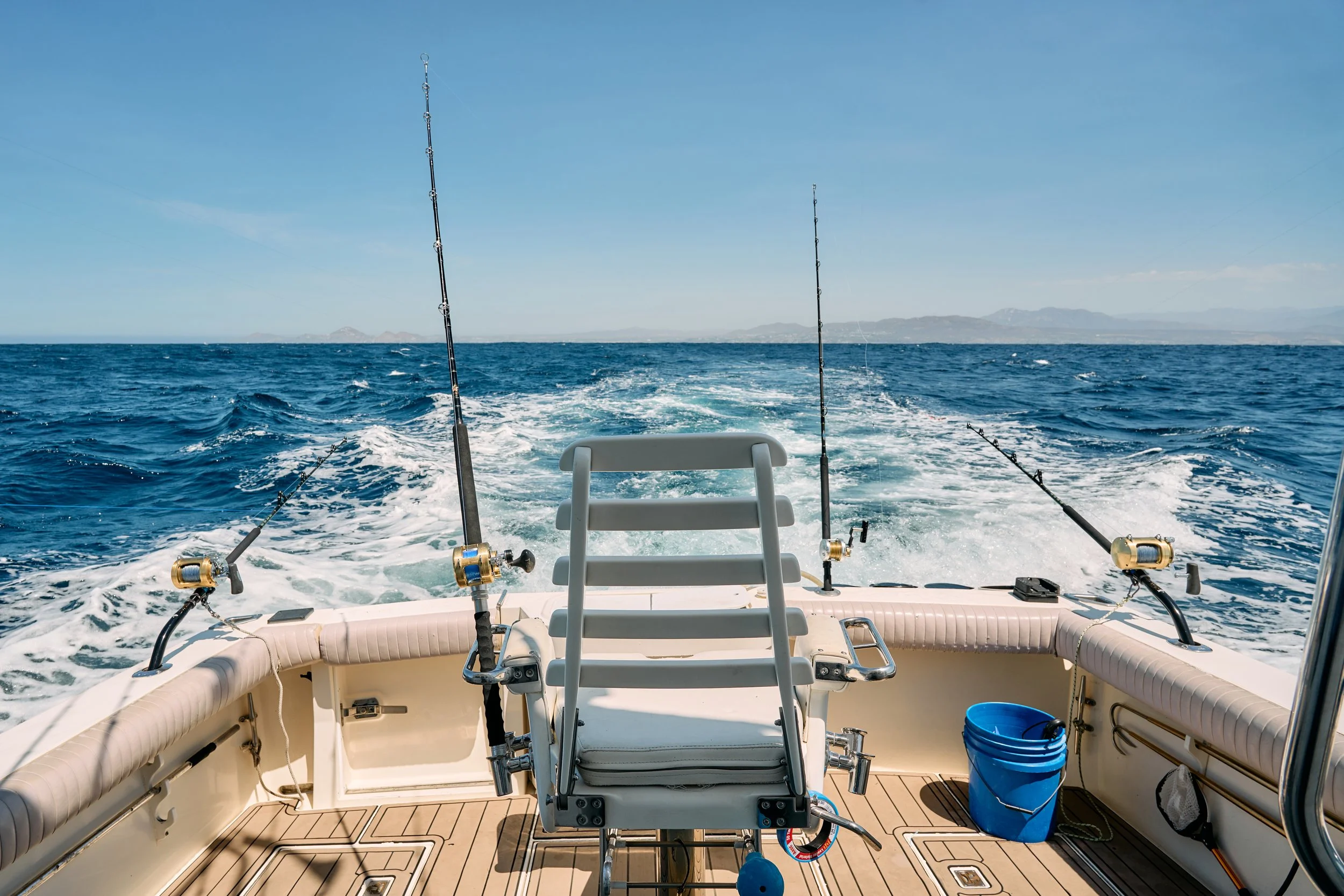 View from the back of a boat on the water with fishing rods mounted on the sides and distant land visible on the horizon under a clear blue sky.