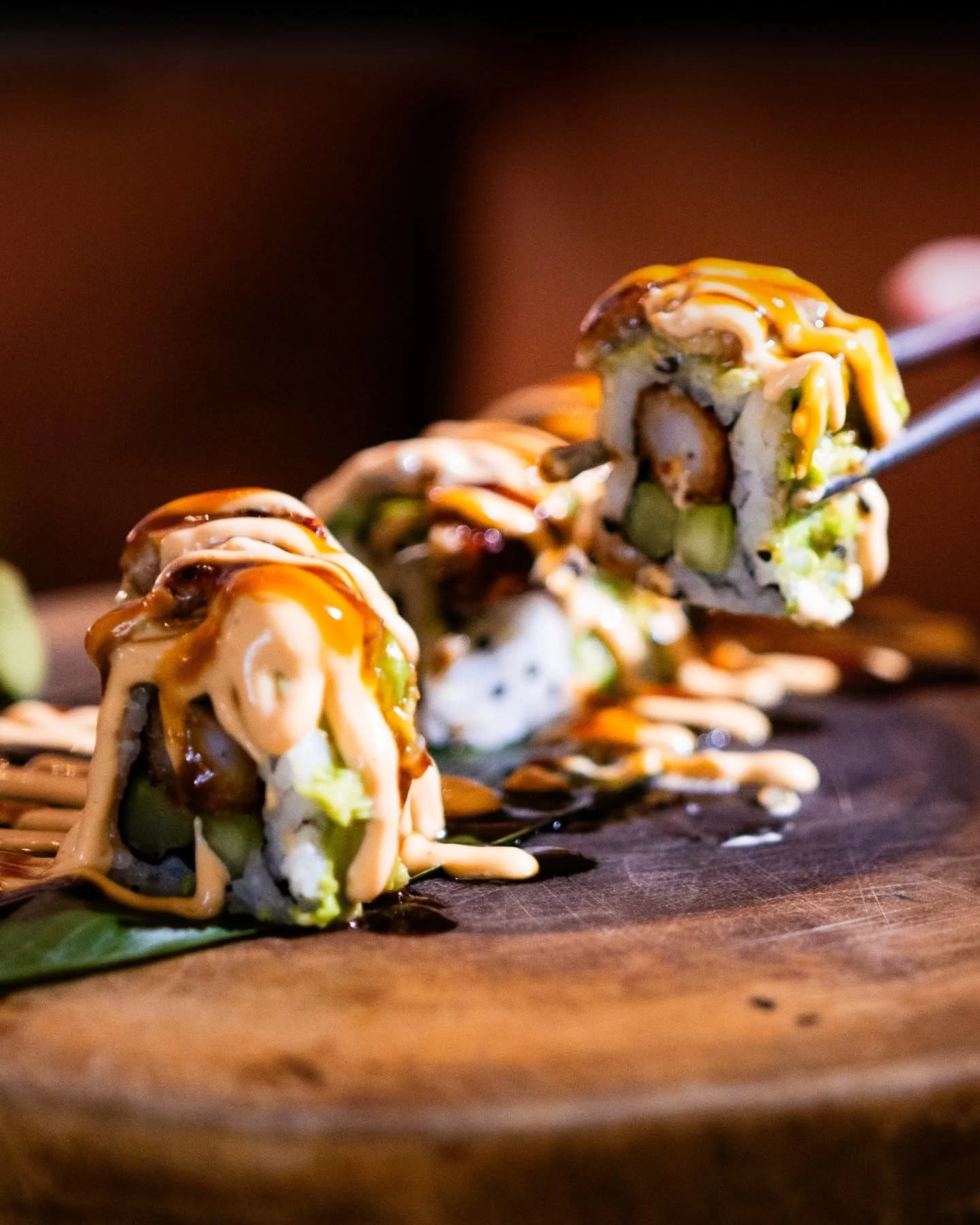 Close-up of sushi rolls topped with sauces, placed on a wooden surface.