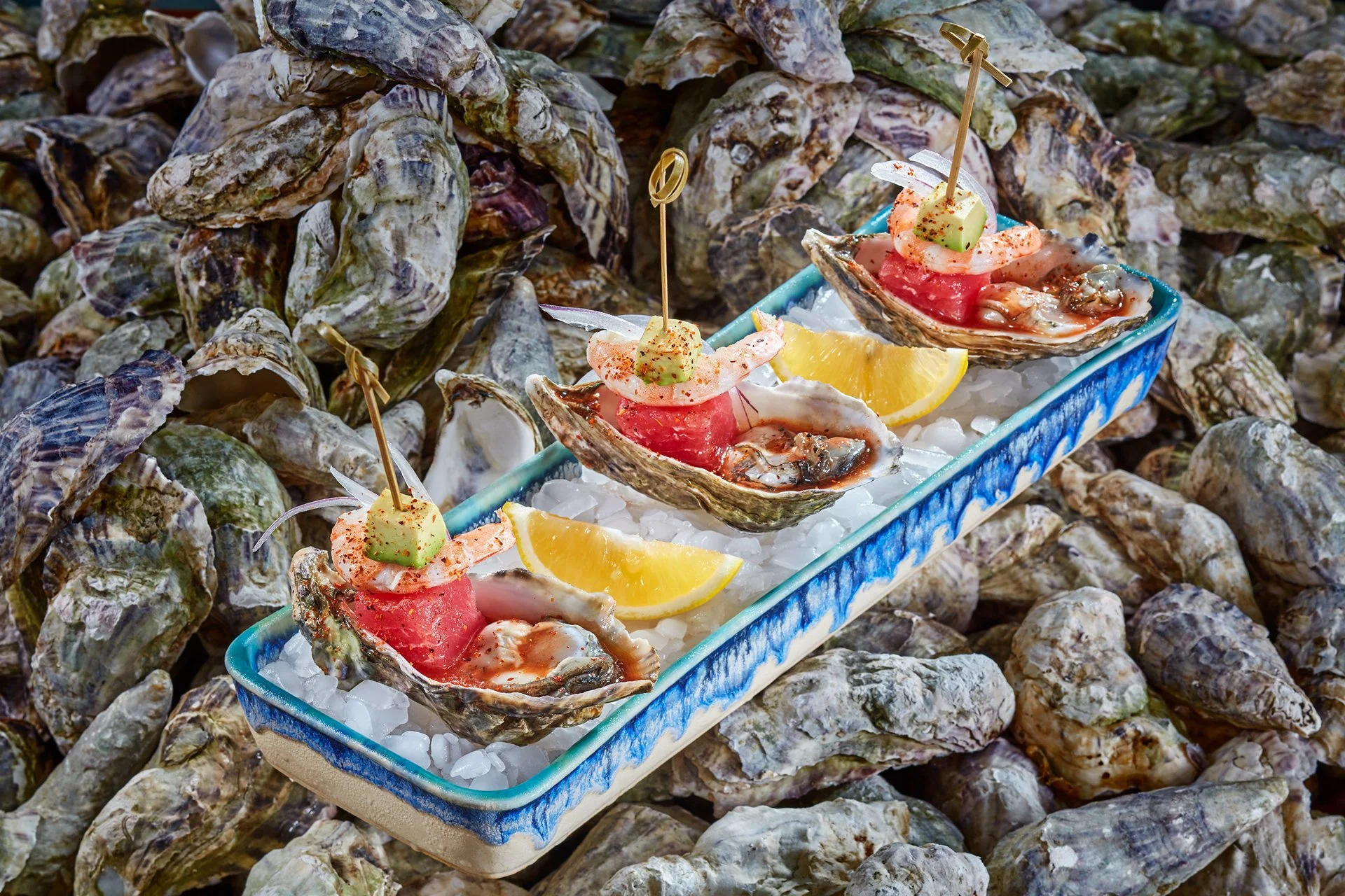 A rectangular serving dish with oysters on a bed of ice, garnished with lemon wedges, slices of cucumber, and a small skewer with seafood and seasoning, surrounded by a pile of oyster shells.