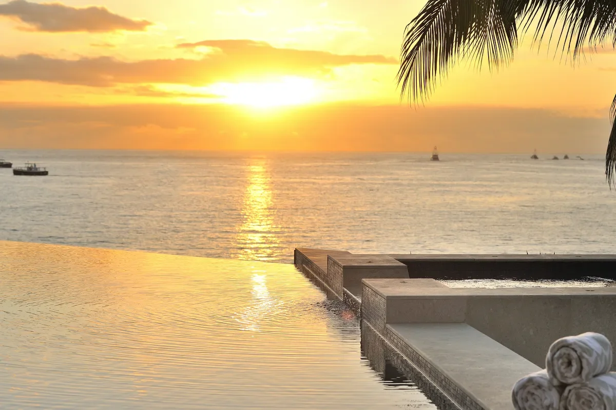 Sunset over the ocean with a pool in the foreground. Palm leaves on the right, several boats on the water, and rolled towels resting on the pool edge.