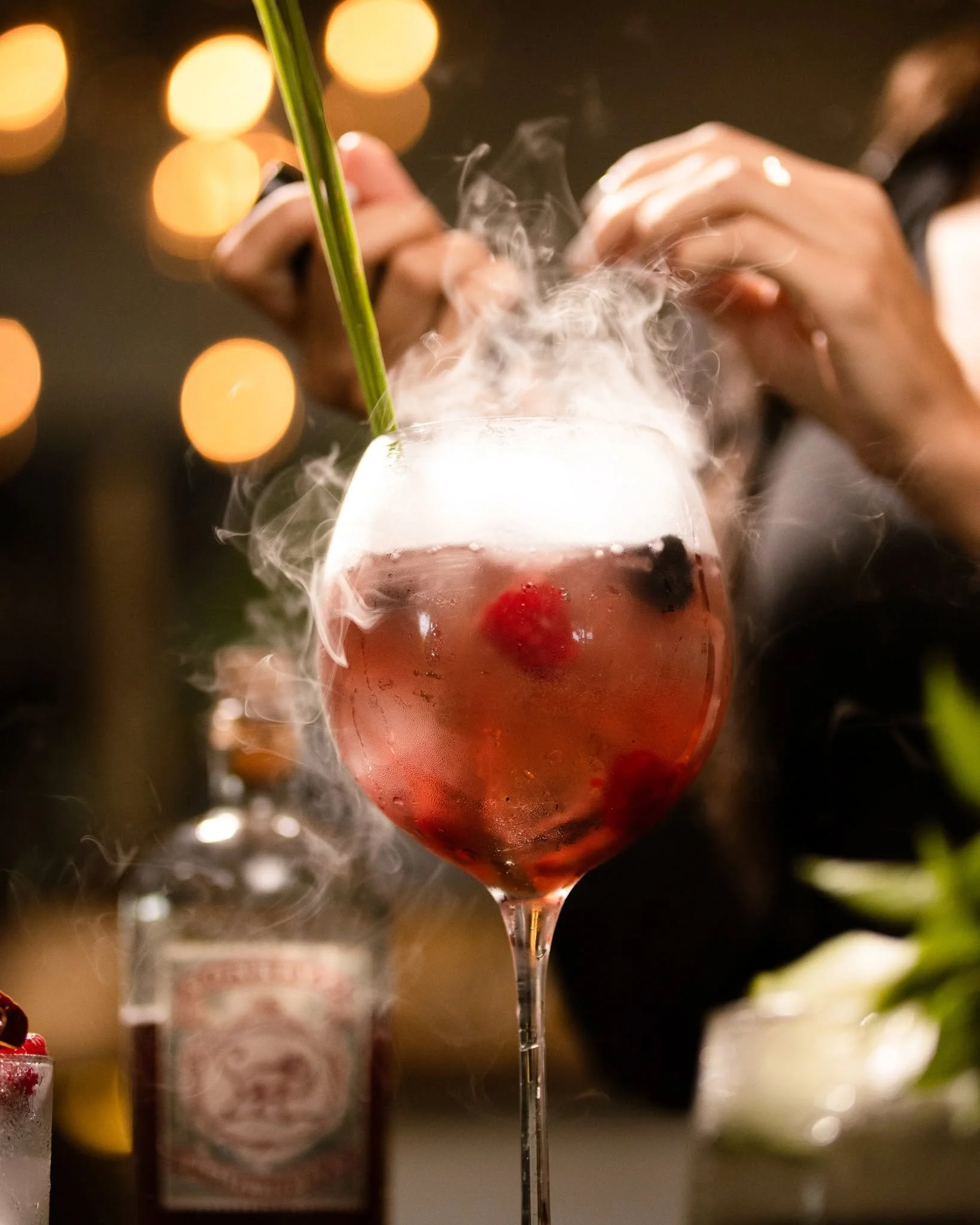 A glass of pink cocktail garnished with a cherry and a green straw, with steam or mist rising from it, against blurred warm bokeh lights in the background.