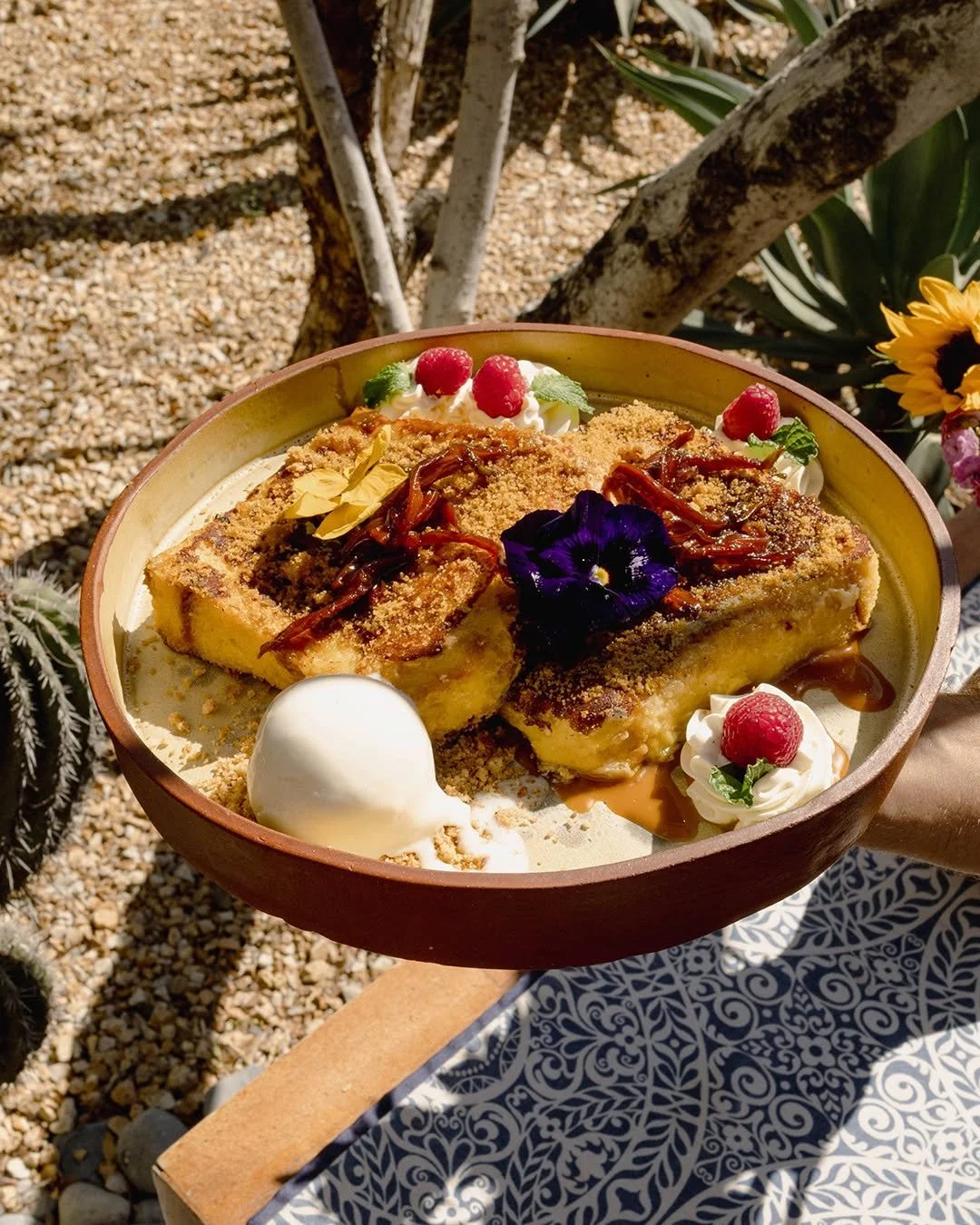 A dessert dish featuring bread pudding topped with caramel sauce, whipped cream, raspberries, mint leaves, edible flower, and a scoop of vanilla ice cream, presented in a bowl outdoors on a decorative table.