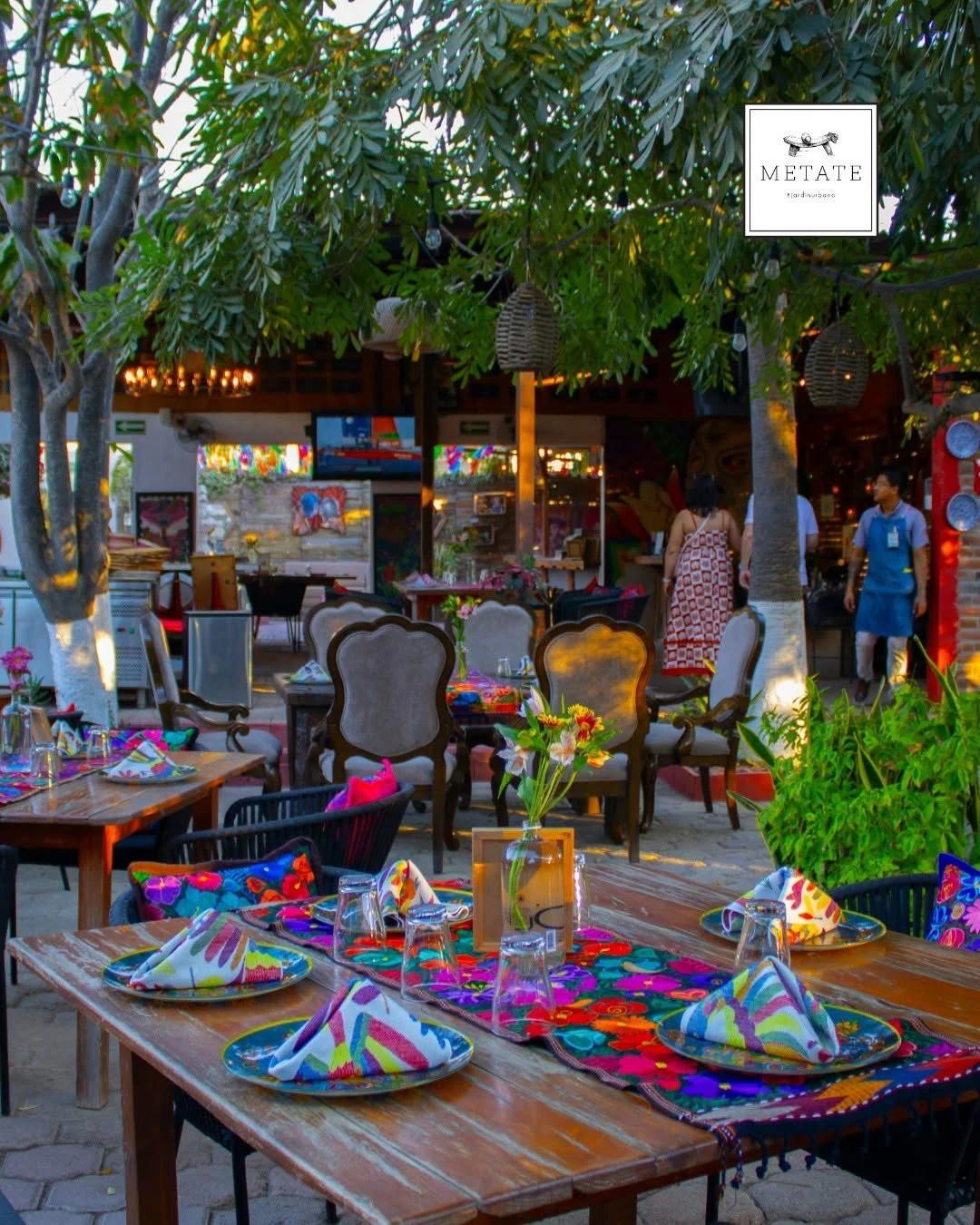 Colorful outdoor restaurant setup with wooden tables, vibrant tablecloths, and neatly folded napkins. The scene is decorated with flowers, green plants, and trees, creating a lively and inviting atmosphere.