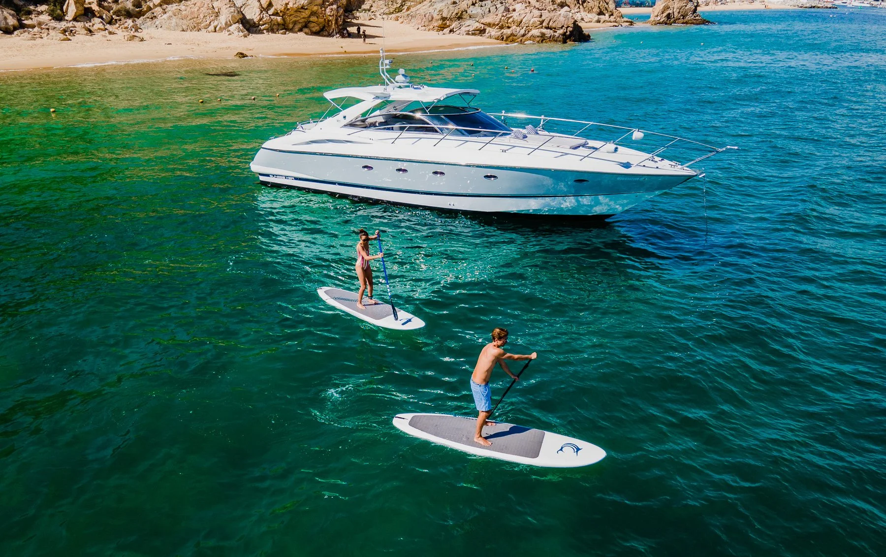 People paddleboarding in turquoise water near a rocky coastline with a large white yacht anchored nearby.