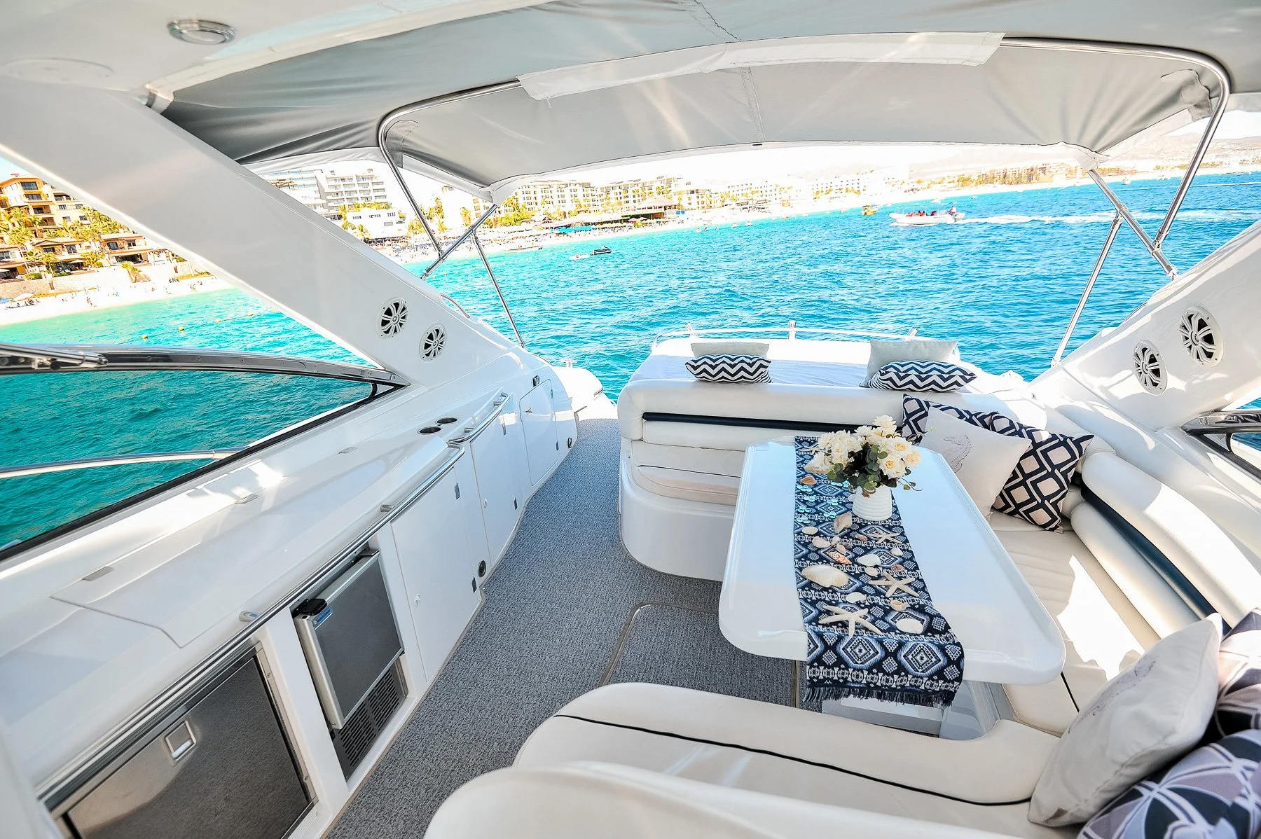 View of the deck area on a yacht with seating, a table adorned with seashells and flowers, and a city and water background under a clear sky.
