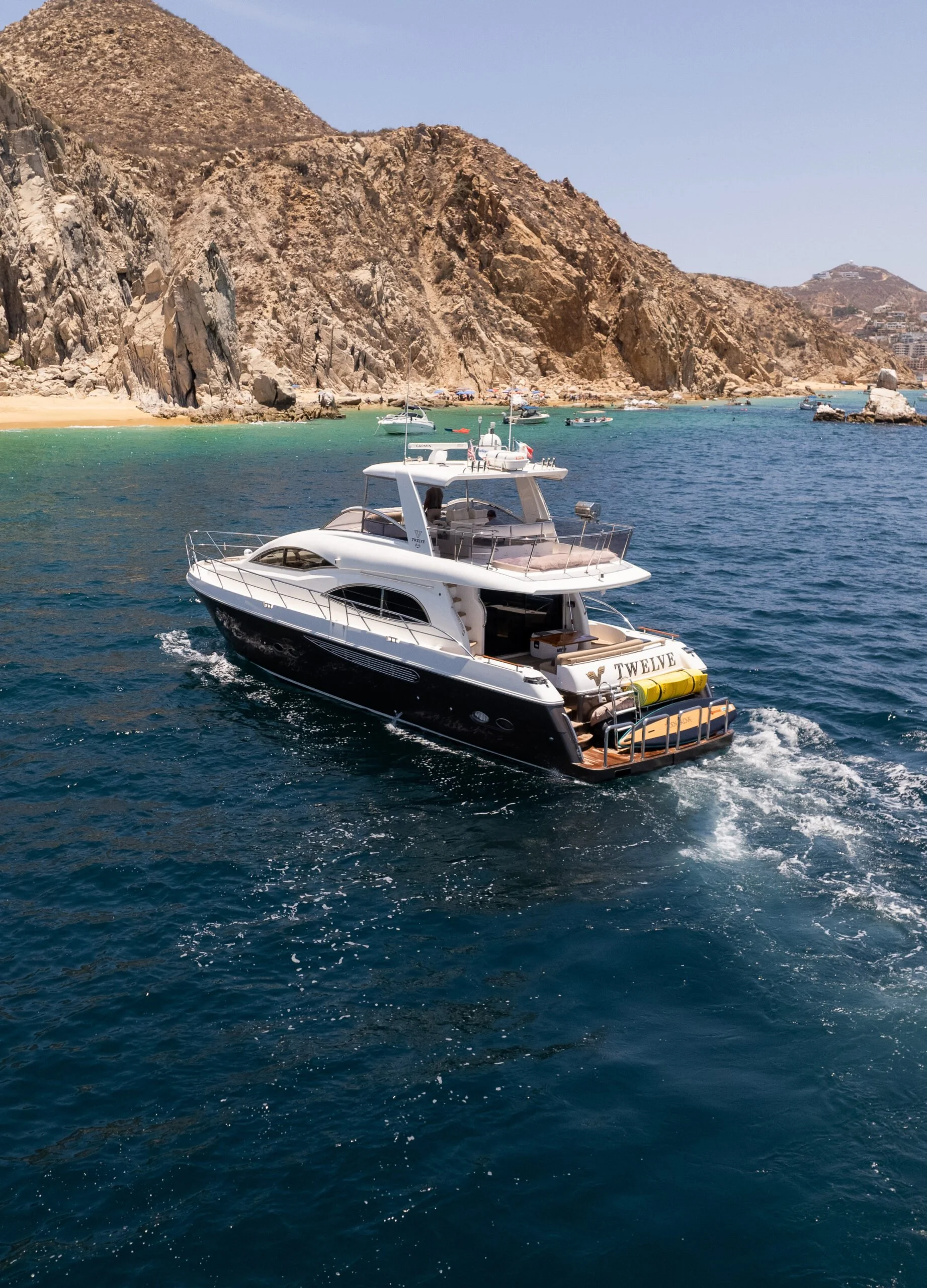 A white yacht named 'Twelve' cruising on clear blue water near a rocky coastline with numerous smaller boats and a sandy beach in the background.
