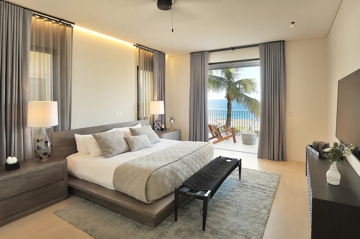 A modern bedroom with a large bed, bedside lamps, and a balcony view of a palm tree and the ocean.