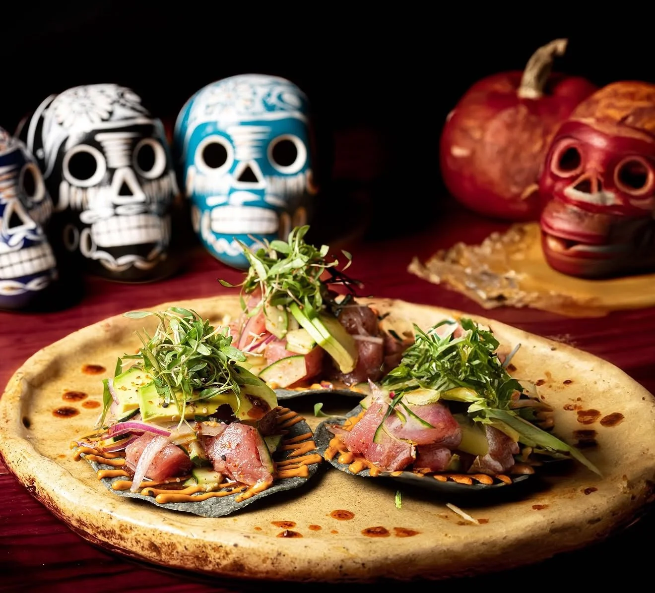 A plate with tacos topped with greens and sauces, with decorative sugar skulls and pumpkins in the background.