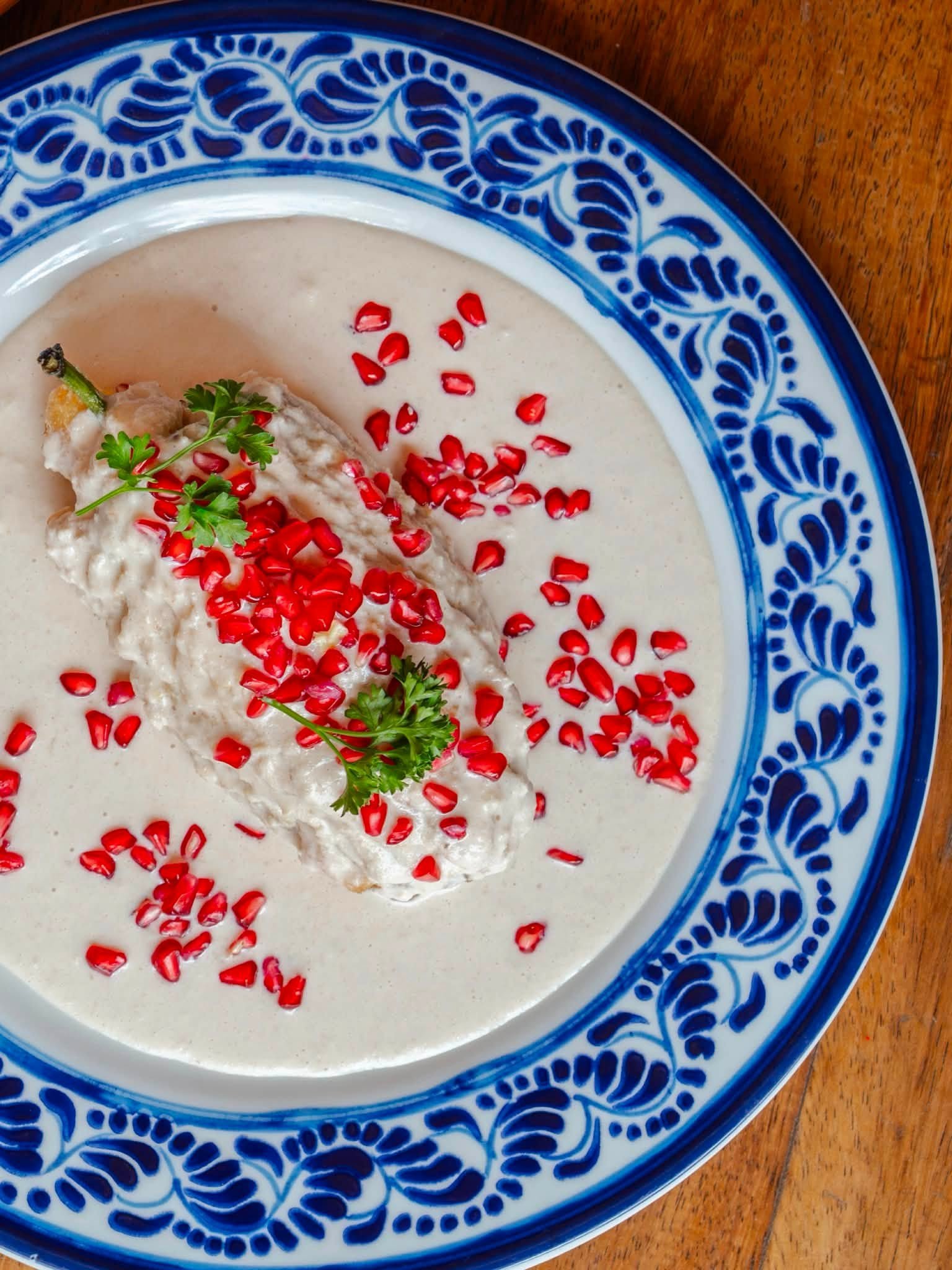 A blue and white ceramic bowl containing a creamy white soup garnished with red pomegranate seeds, a sprig of cilantro, and a piece of chicken leg, placed on a wooden surface.