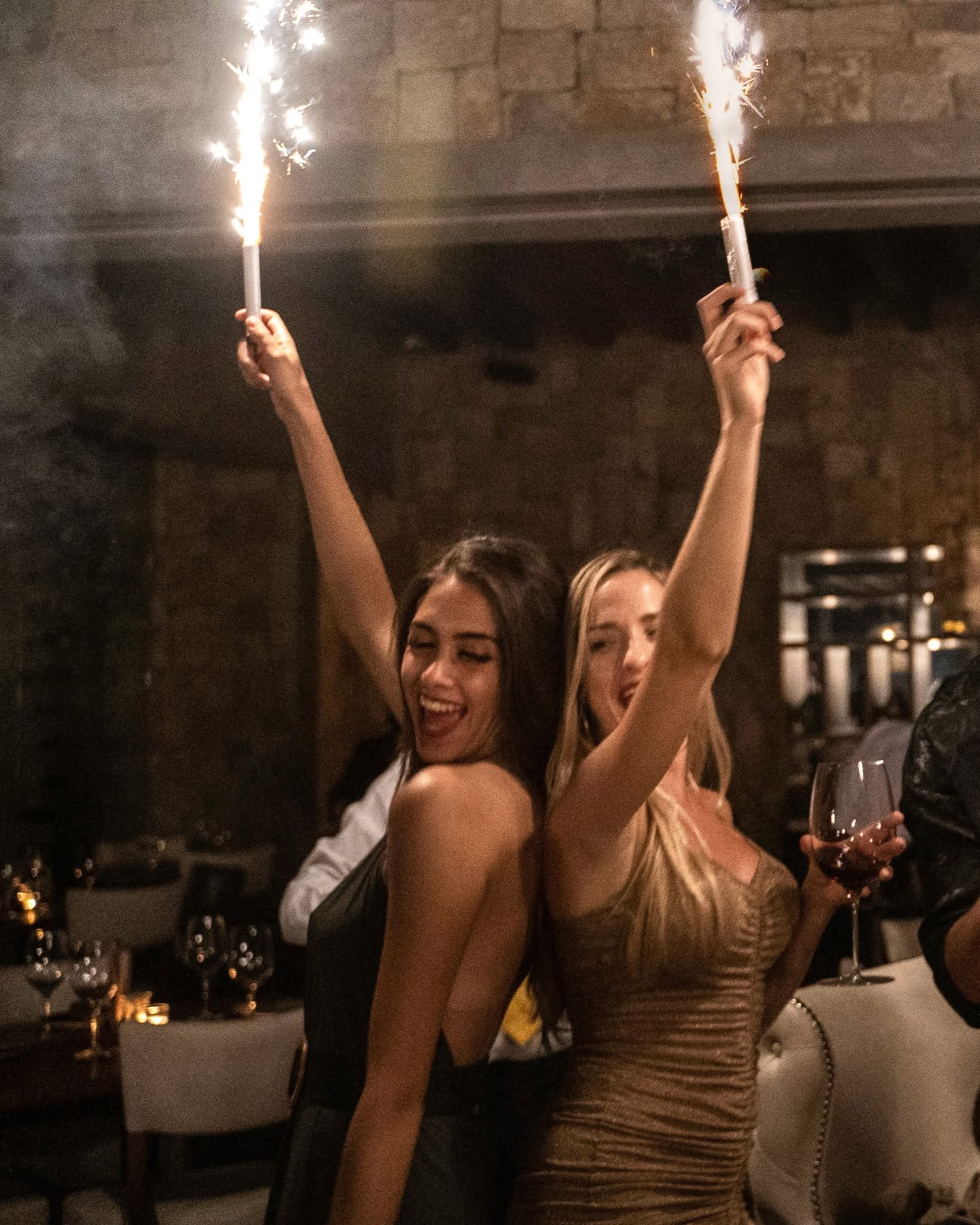 Two women holding sparklers at an indoor celebration, with glasses of red wine on the table behind them.