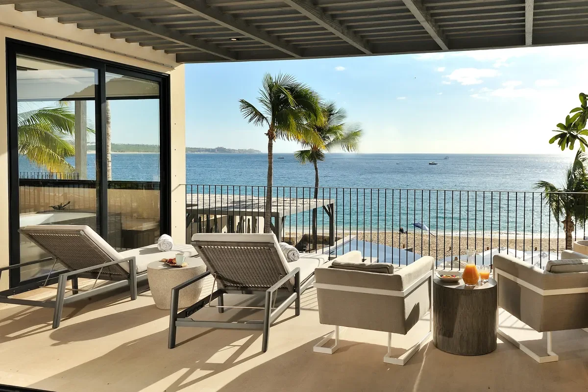 Beachfront patio with lounge chairs, small tables, and a view of the ocean, palm trees, and clear sky